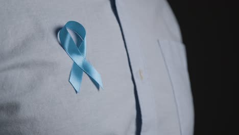 Close-Up-Of-Man-Putting-On-Blue-Ribbon-Badge-Symbolizing-Awareness-Of-Men's-Health-And-Cancer-Onto-Shirt